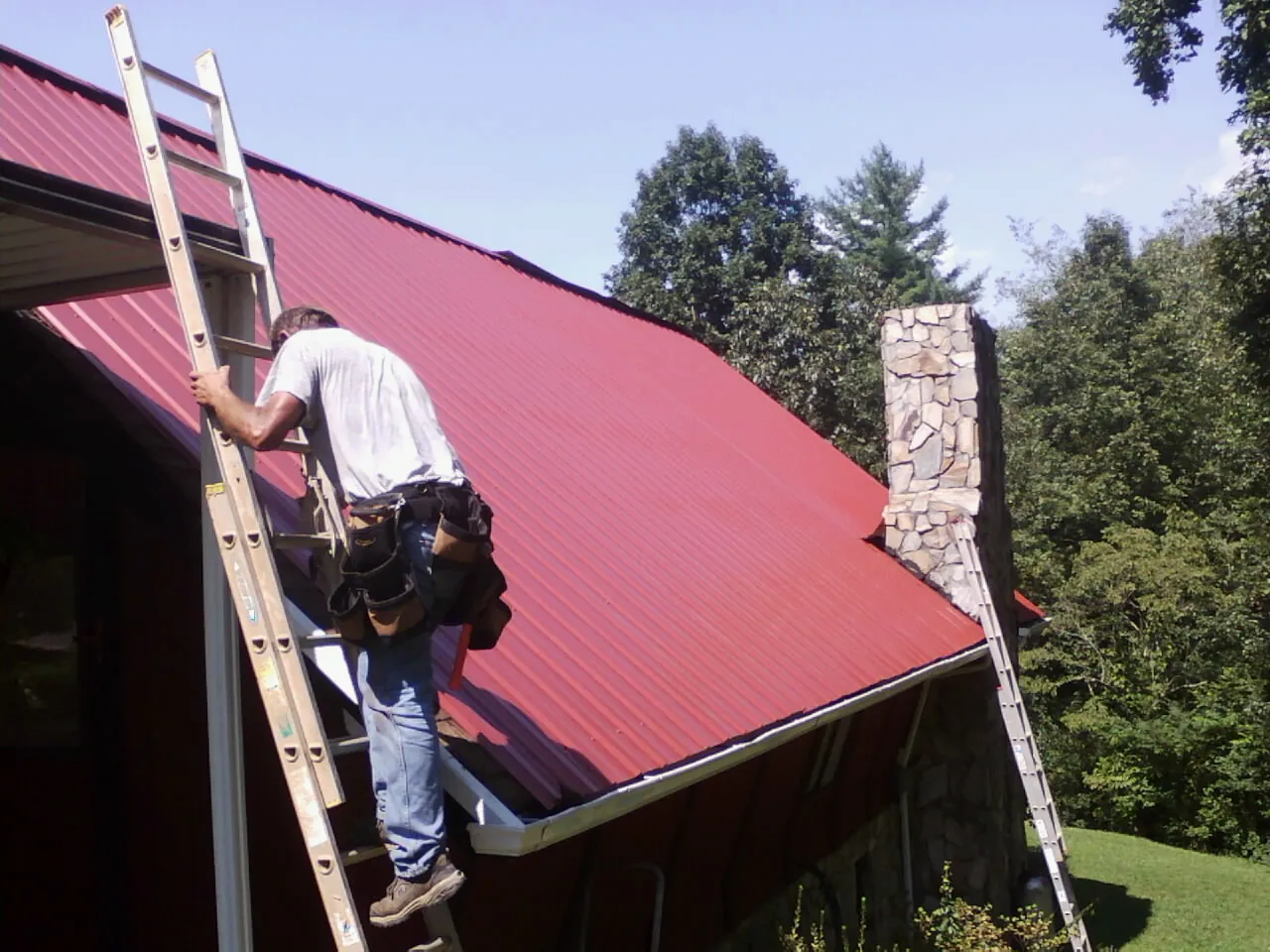 Roofer on a ladder beside a red metal roof and stone chimney — Soffit & Fascia Repair by Roofing Brothers in Silsbee
